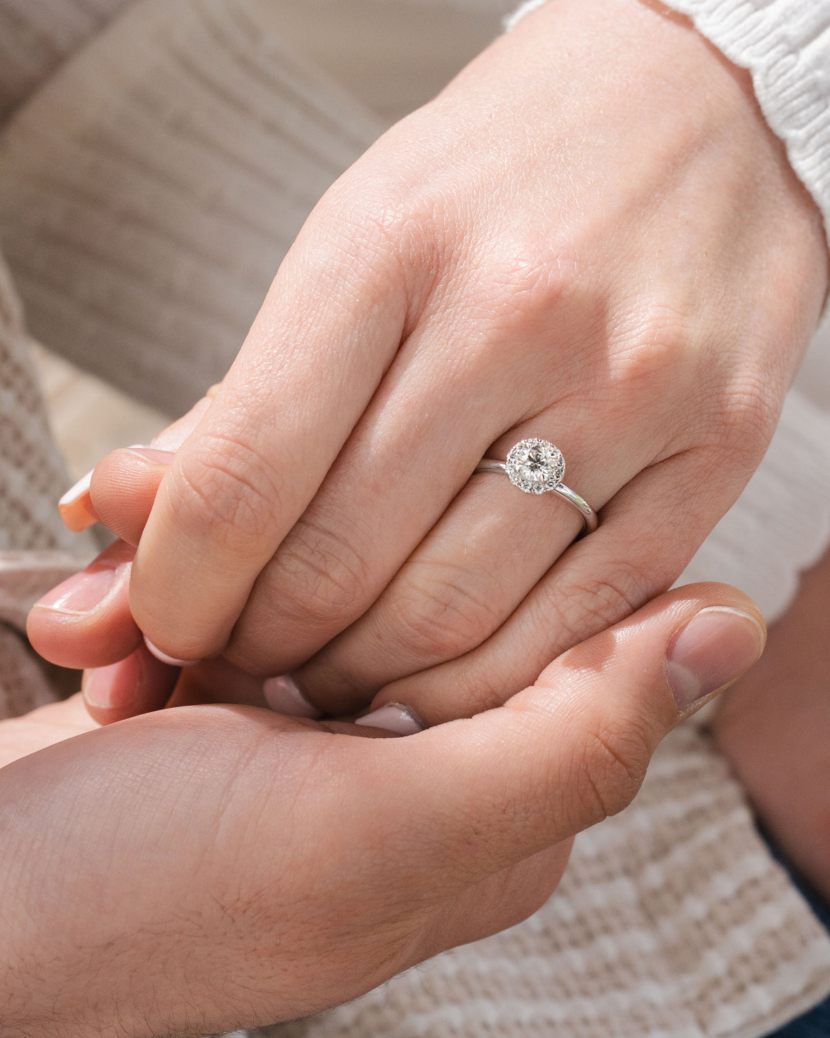 A close-up of a silver halo engagement ring with a round-cut clear gemstone worn on a woman's hand, which is being held gently by a man's hand. The background shows soft beige knit clothing.