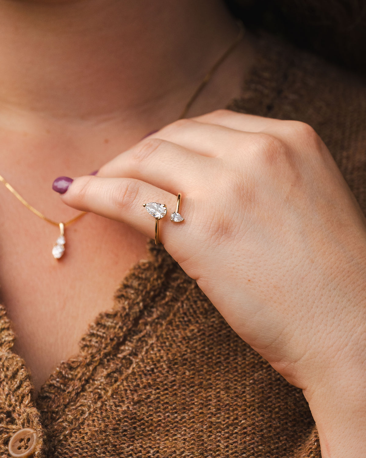 A close-up lifestyle shot of a person's hand wearing a delicate gold ring with a clear marquise-cut gemstone and a small pearl accent. The person is also wearing a gold necklace with a white pearl drop, set against a brown knit cardigan.