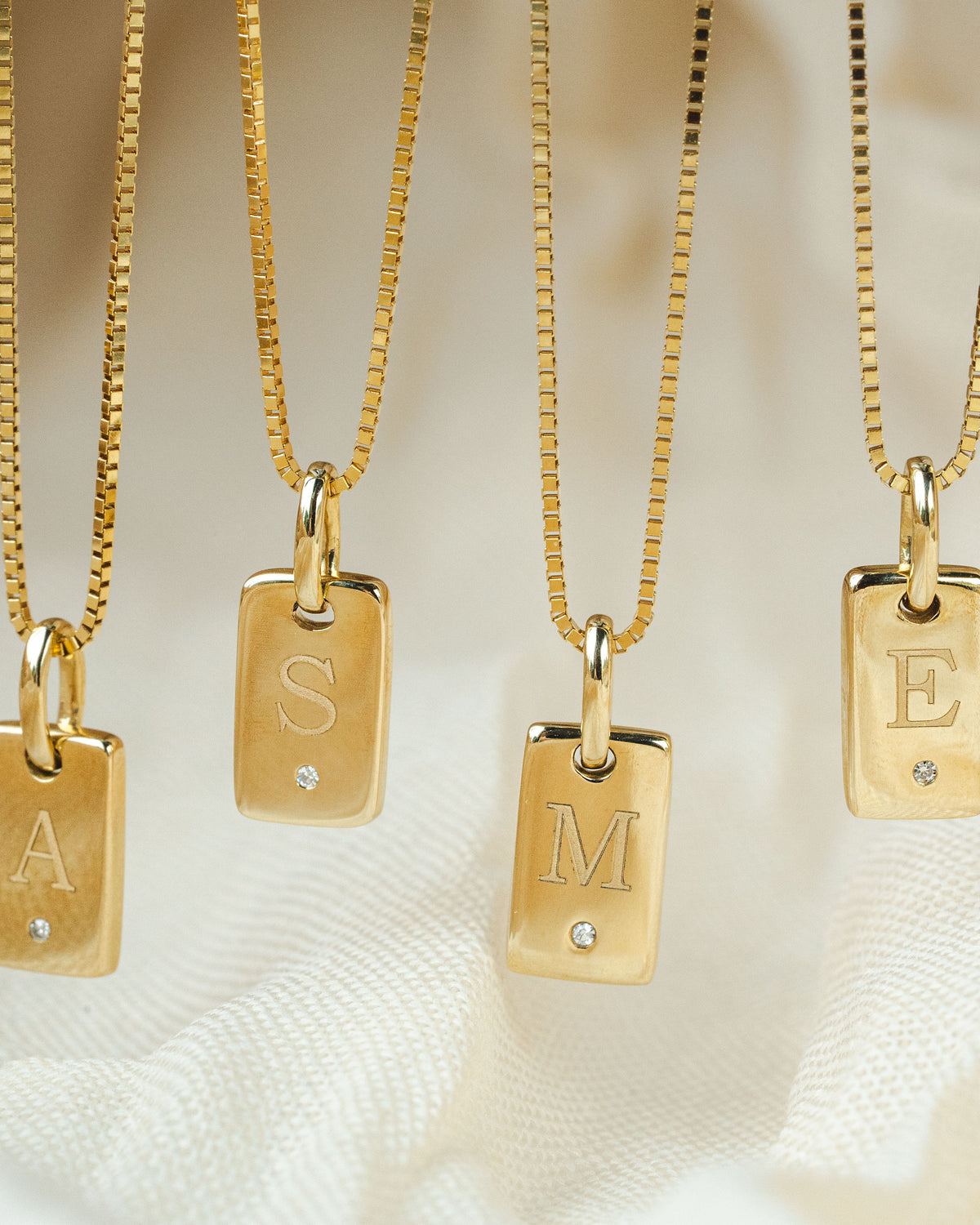 Four gold initial necklaces with rectangular engraved pendants hanging against soft white fabric. The pendants are labeled A, S, M, and E, each with a small clear stone.