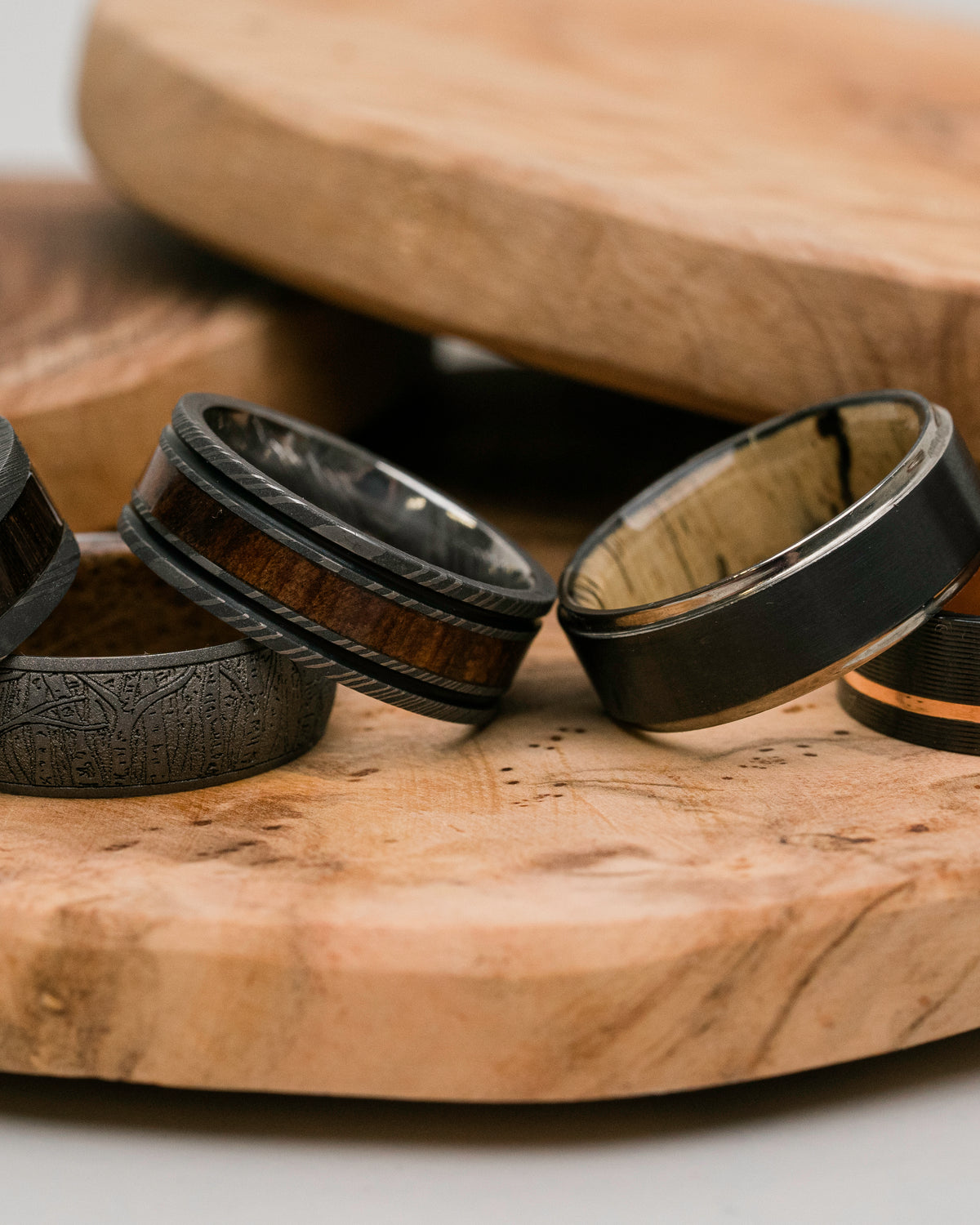 Four wooden wedding bands with different inlay patterns and colors, displayed on a natural wood serving board.