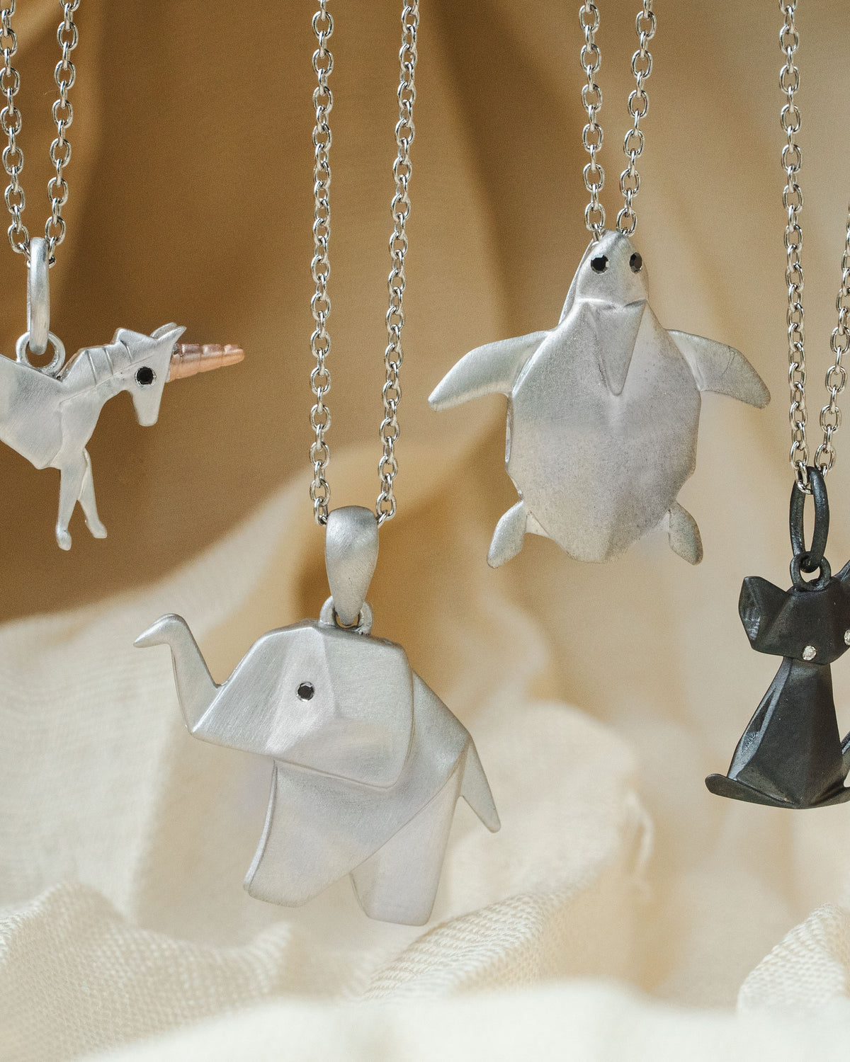 Four origami-style animal pendant necklaces hanging from silver chains against a soft beige fabric backdrop. The pendants include a unicorn, turtle, elephant, and cat, with three in silver and one in dark gray.