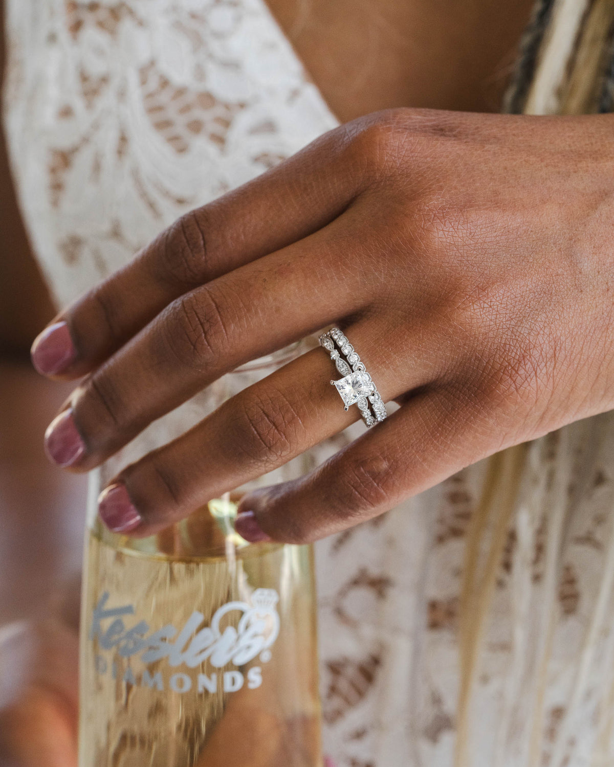 A close-up of a hand wearing a silver diamond engagement ring, holding a bottle of sparkling wine. The hand has pink manicured nails, and the person is wearing a white lace dress.