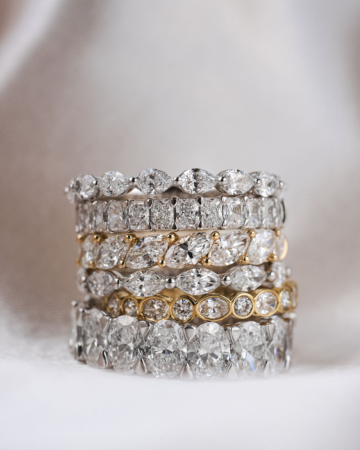 A stack of five diamond eternity rings in silver and yellow gold settings, displayed on a soft white fabric background.