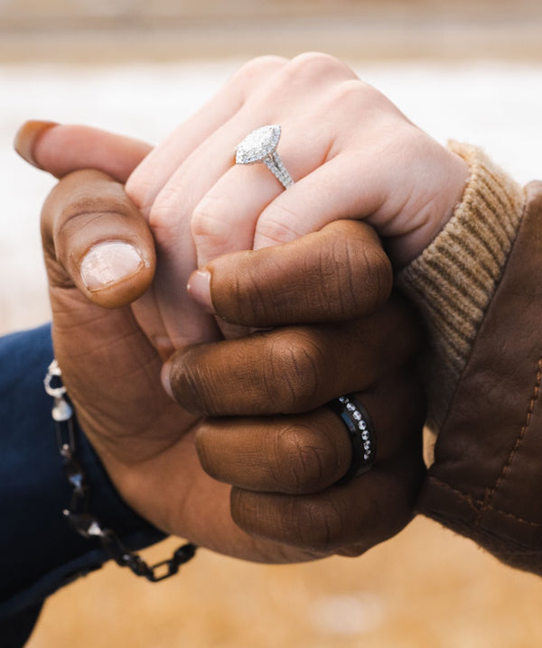 A close-up of two hands holding each other, with a silver diamond engagement ring on the lighter-skinned hand and a black diamond band on the darker-skinned hand.