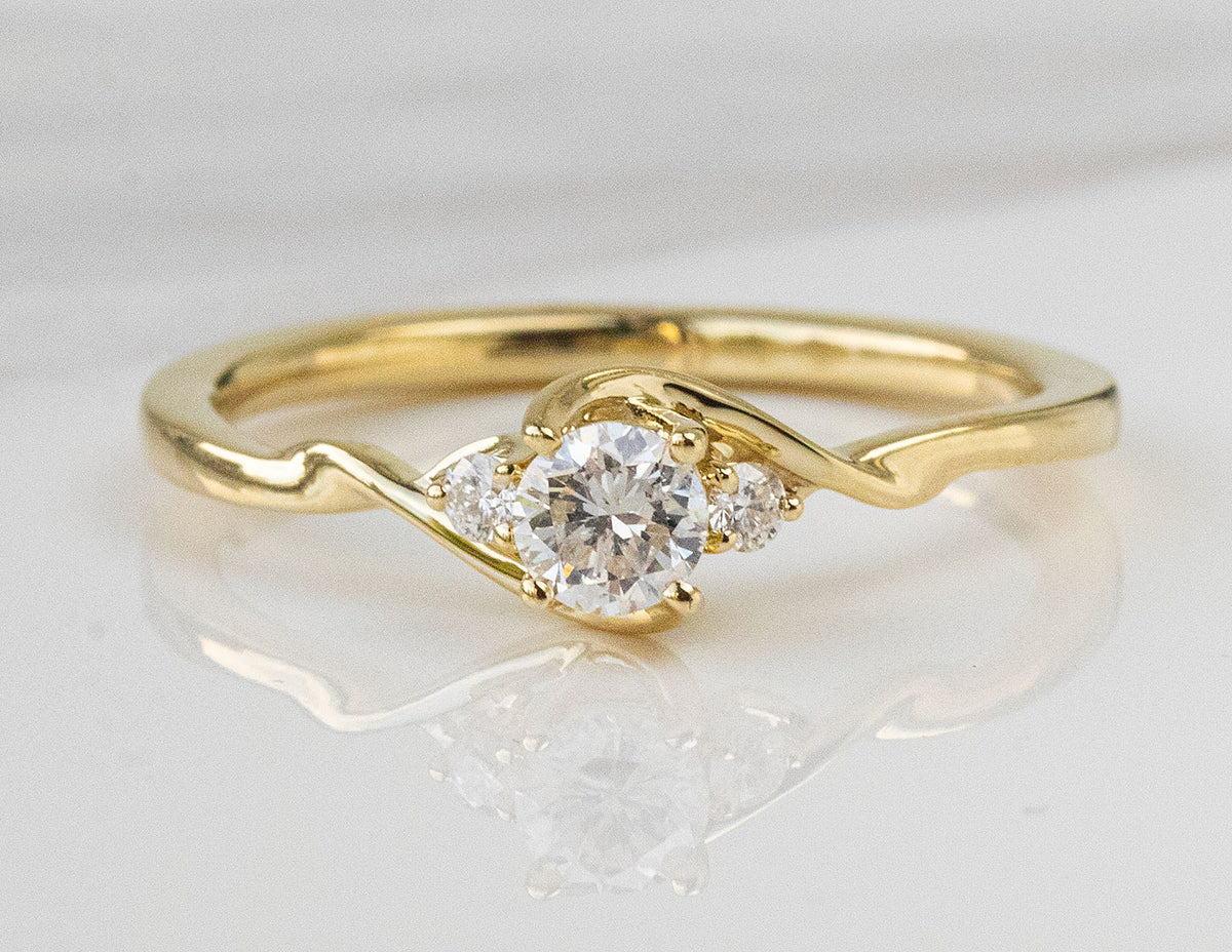 A yellow gold solitaire engagement ring with a round-cut center stone and small side stones, displayed on a reflective white surface.