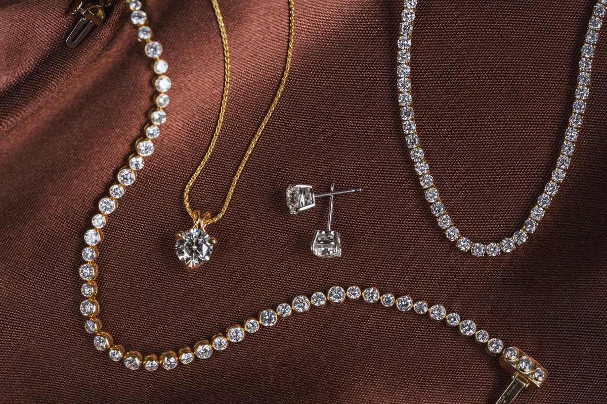 A luxury jewelry set featuring a gold pendant necklace with a large clear gemstone, a silver stud earring, and multiple diamond-studded bracelets and necklaces, arranged on a rich brown satin fabric.