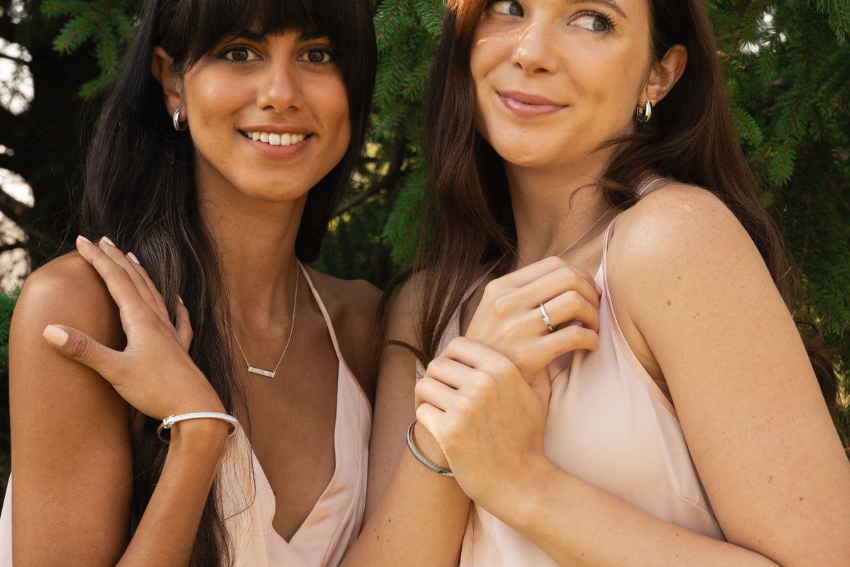 Two women posing outdoors in front of green foliage, wearing matching pale blush pink satin camisole dresses and displaying silver jewelry, including rings and bracelets.
