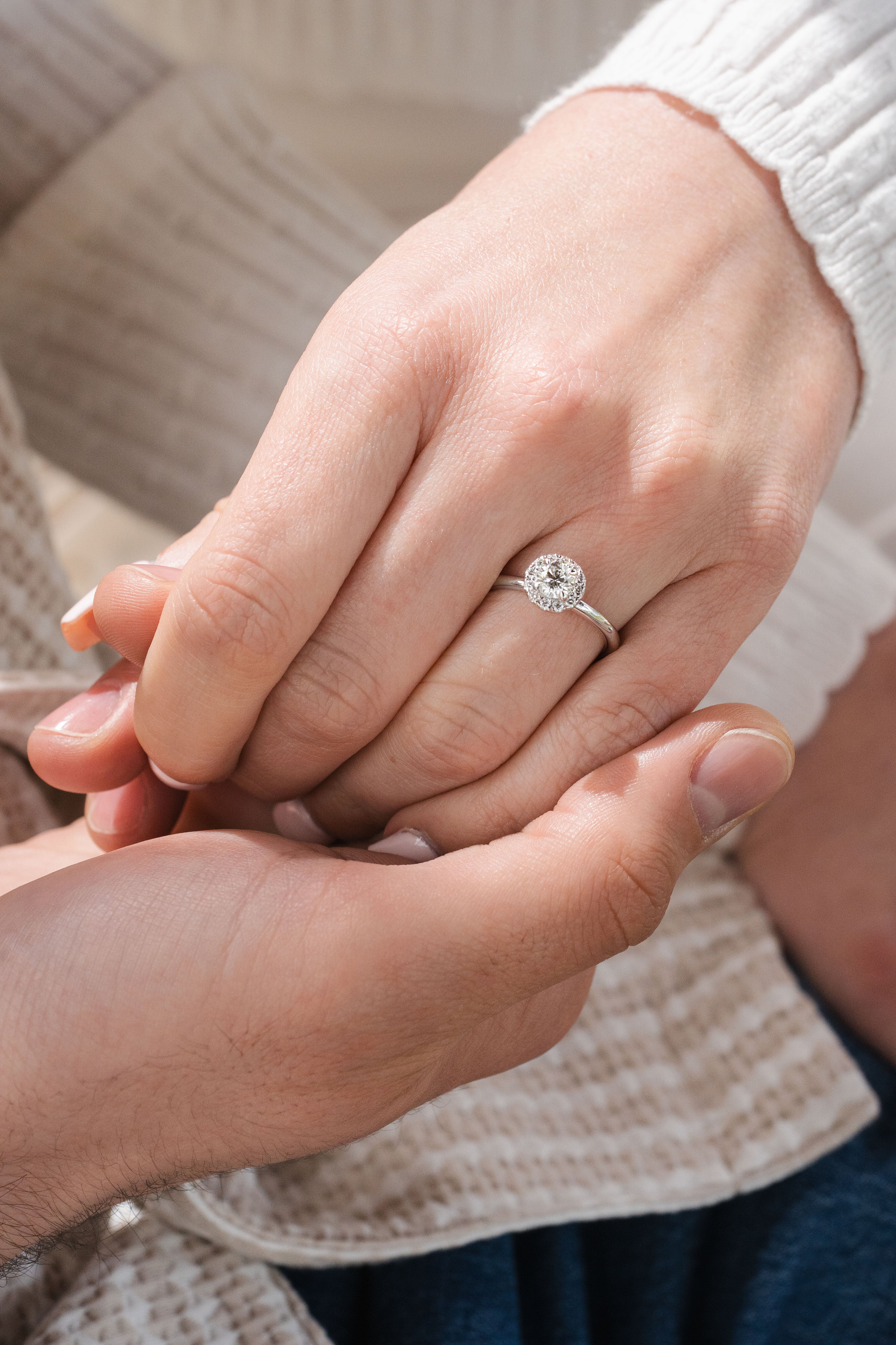 A close-up of a silver halo engagement ring with a round-cut clear gemstone worn on a woman's hand, which is being held gently by a man's hand. The background shows soft beige knit clothing.