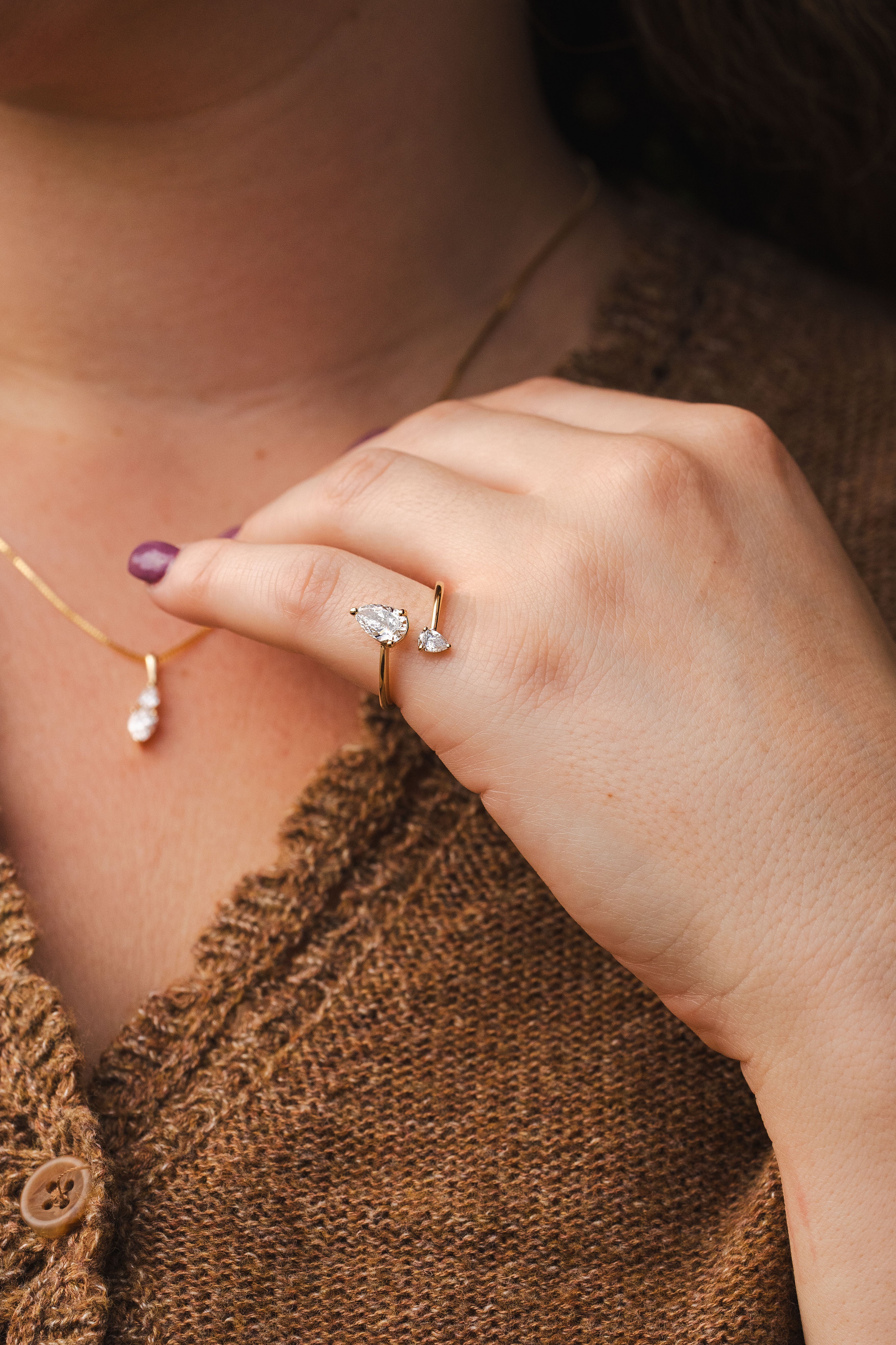 A close-up lifestyle shot of a person's hand wearing a delicate gold ring with a clear marquise-cut gemstone and a small pearl accent. The person is also wearing a gold necklace with a white pearl drop, set against a brown knit cardigan.