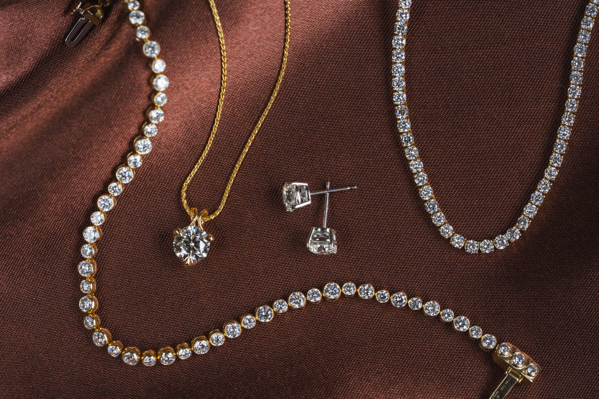 A luxury jewelry set featuring a gold chain necklace with a clear round-cut diamond pendant, a silver stud earring, and multiple diamond-studded tennis necklaces in gold and silver, arranged on a brown satin fabric background.