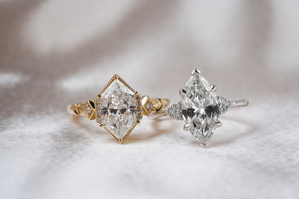 Two diamond rings on a soft white fabric background, one in yellow gold and one in silver, each featuring a marquise-cut diamond with smaller accent diamonds.