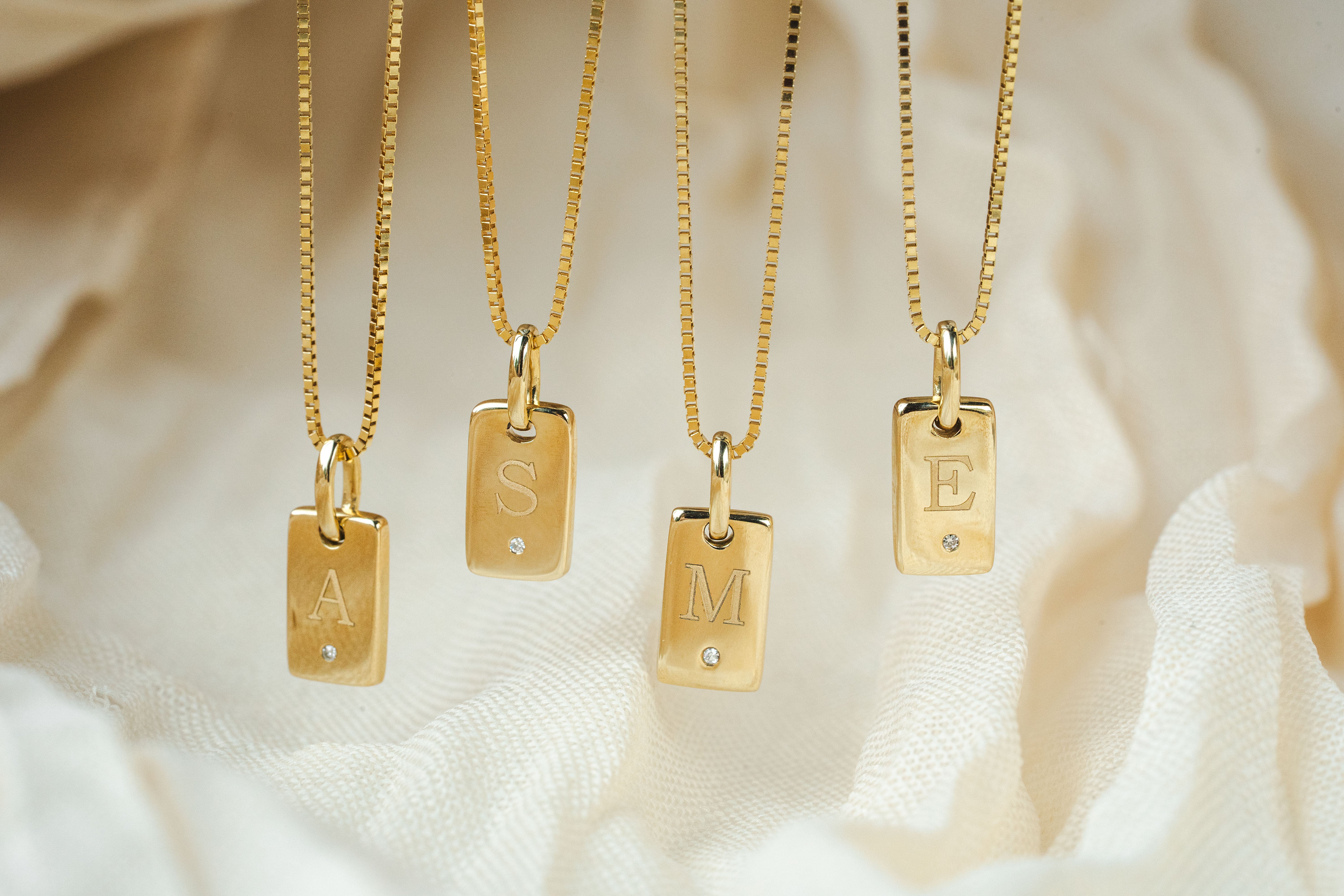 Four gold initial necklaces with rectangular engraved pendants hanging against soft white fabric. The pendants are labeled A, S, M, and E, each with a small clear stone.