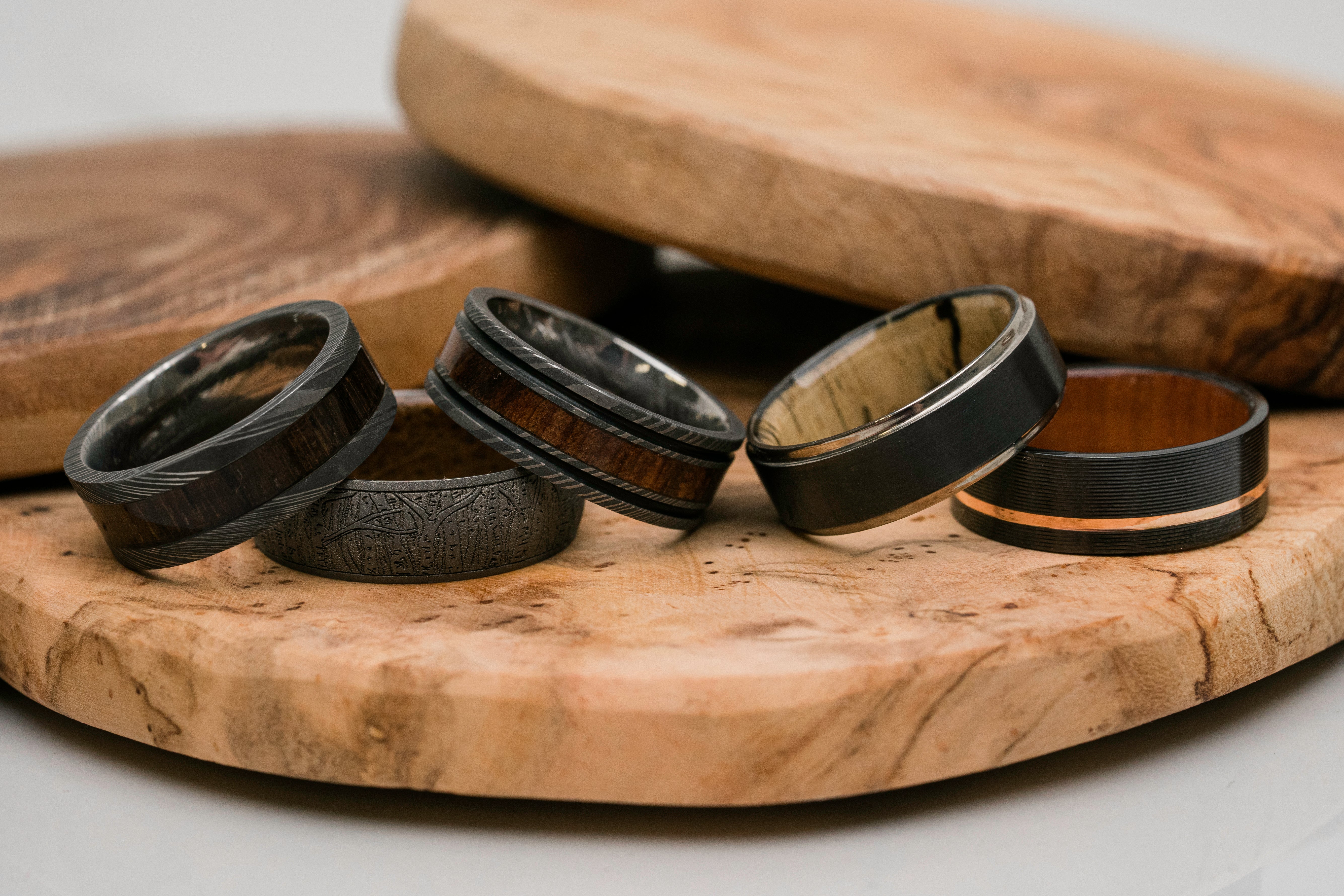Four wooden wedding bands with different inlay patterns and colors, displayed on a natural wood serving board.