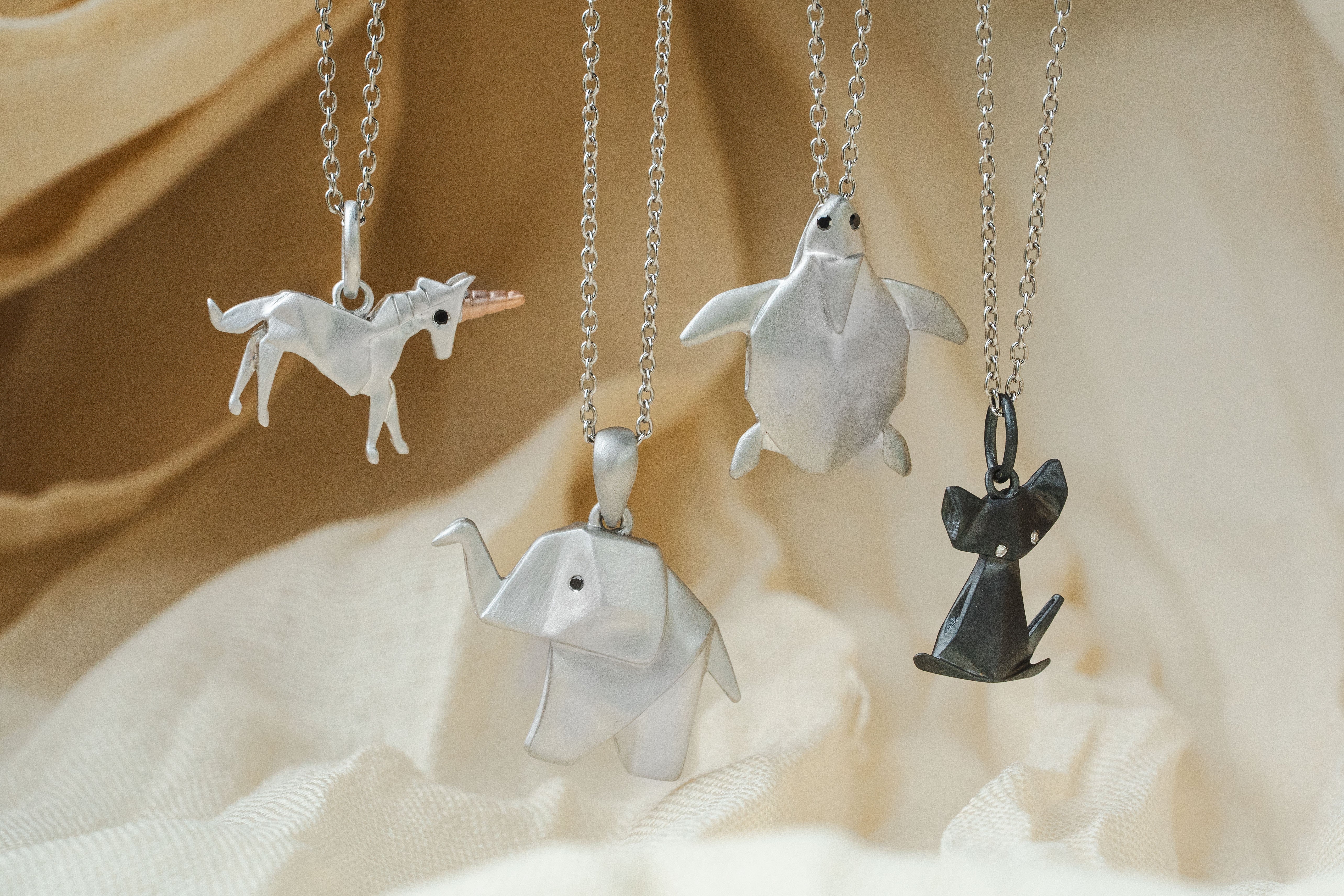 Four origami-style animal pendant necklaces hanging from silver chains against a soft beige fabric backdrop. The pendants include a unicorn, turtle, elephant, and cat, with three in silver and one in dark gray.