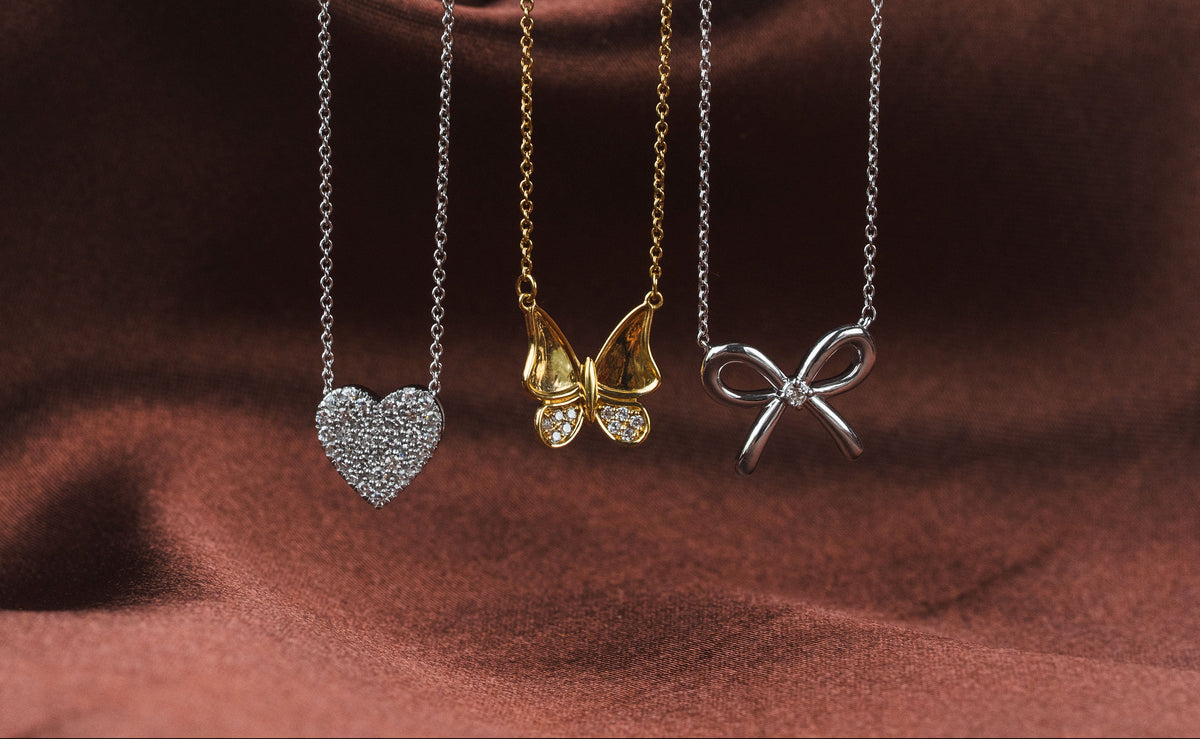 Three delicate necklaces with butterfly and geometric pendants displayed against a draped dark brown satin fabric. The necklaces feature silver and gold finishes with sparkling clear stones.