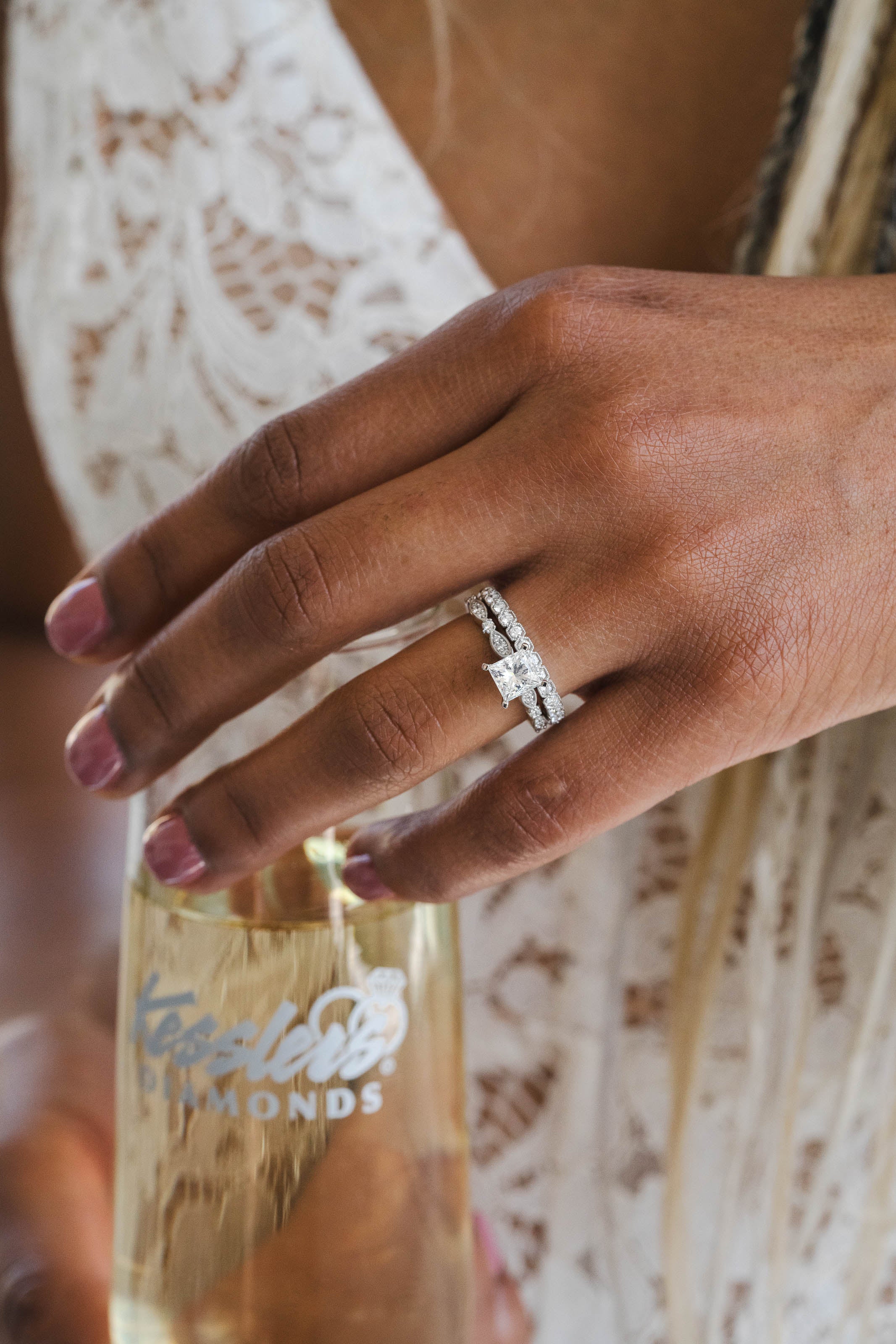 A close-up of a hand wearing a silver diamond engagement ring, holding a bottle of sparkling wine. The hand has pink manicured nails, and the person is wearing a white lace dress.