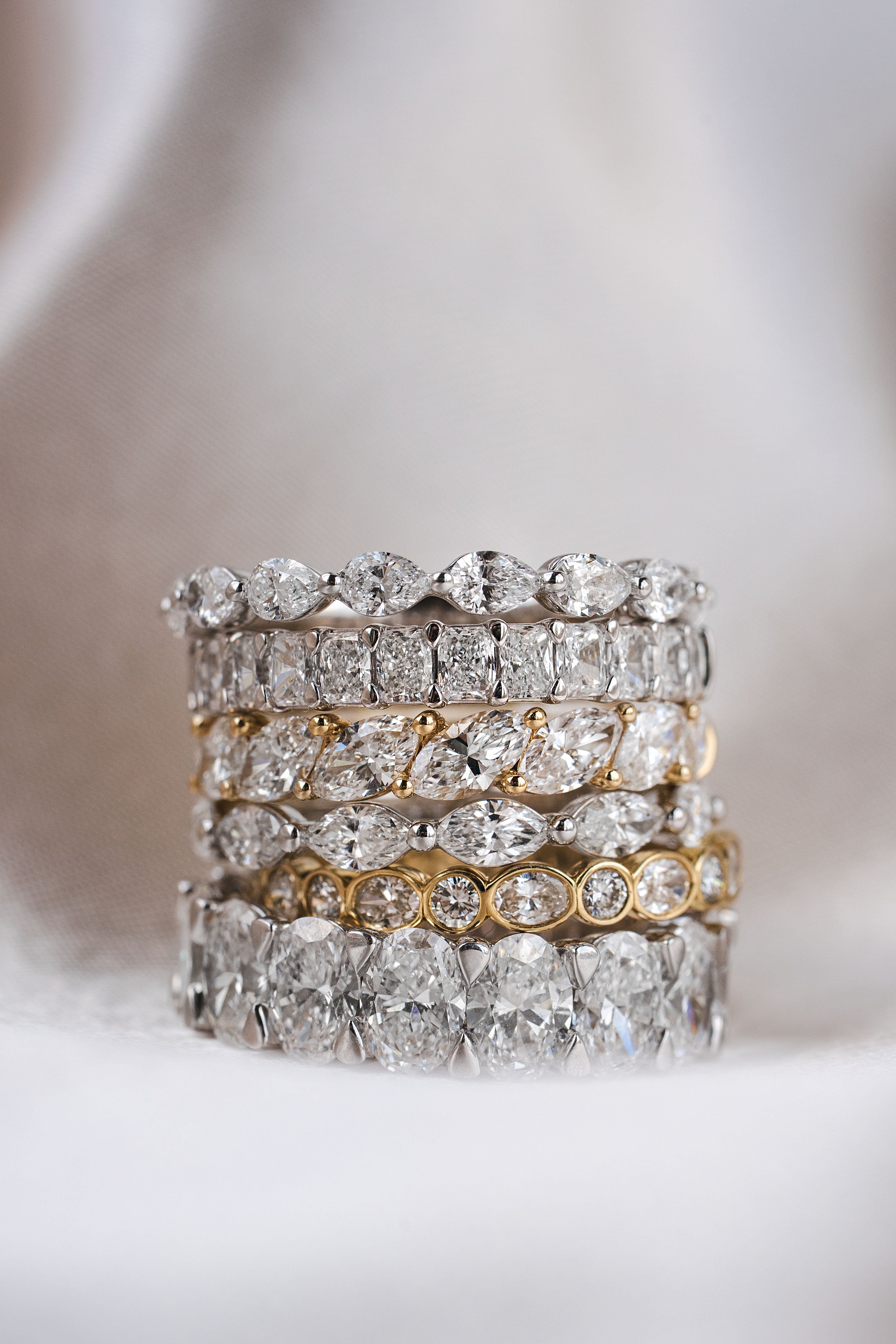 A stack of five diamond eternity rings in silver and yellow gold settings, displayed on a soft white fabric background.