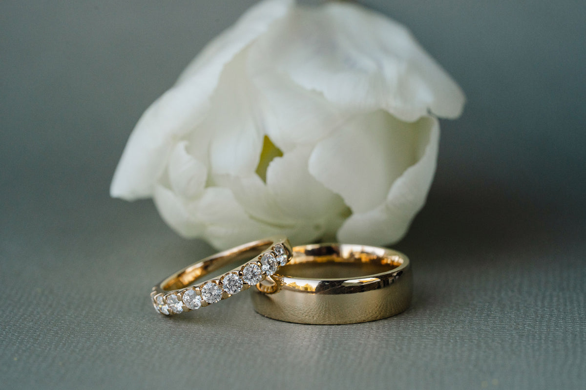 Two gold wedding rings, one with a row of clear pavé stones and the other plain, resting in front of a white flower on a gray textured surface.